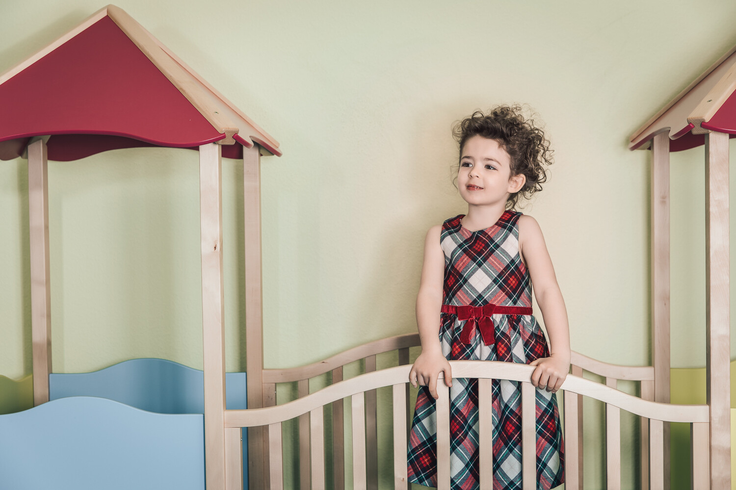 Ein Mädchen in einem rot-weiss karierten Kleid und braunen Locken steht zwischen zwei Türmen und blickt in die Weite. Das Dach der beiden Türme ist rot, während die Wände der Türme blau sind. Die Wand hinter dem Mädchen ist gelb. Die Kindervilla gehört zu dem Wellnesshotel Grand Resort Bad Ragaz, einem Superior Luxushotel in der Schweiz.