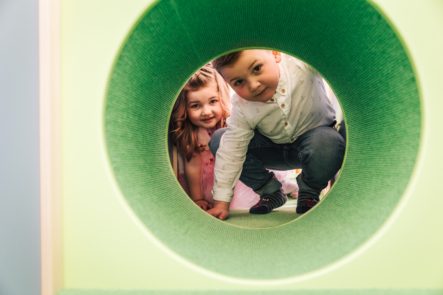 Zwei Kinder, ein Junge und ein Mädchen, schauen durch eine grüne Röhre in die Kamera rein. Die Röhre befindet sich in der Kindervilla. Diese gehört zum Wellnesshotel Grand Resort Bad Ragaz, einem Superior Luxushotel in der Schweiz.
