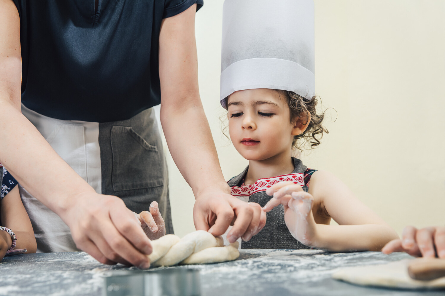 Die Betreuerin hilft dem Mädchen mit braunen Locken einen kleinen Hefezopf zu formen. Der Tisch und die Hände der beiden sind voller Mehl. Das kleine Mädchen trägt eine Schürze und einen grossen, weissen Kochhut. Sie befinden sich in der Kindervilla, die zu dem Wellnesshotel Grand Resort Bad Ragaz gehört, einem Superior Luxushotel in der Schweiz.