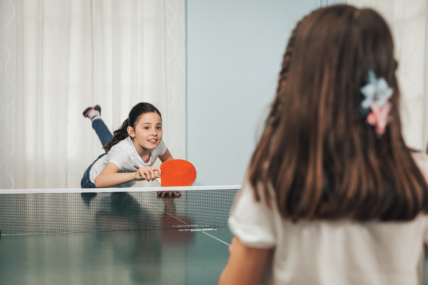 Ein Mädchen mit dunklen Haaren spielt mit ihrer Freundin Tischtennis. Sie hat soeben den Ball geschossen und liegt auf der Tischtennisplatte. Hinter ihr befindet sich ein weisser Vorhang. Die Kindervilla befindet sich in dem Wellnessresort Grand Resort Bad Ragaz, einem Superior Luxushotel in der Schweiz.