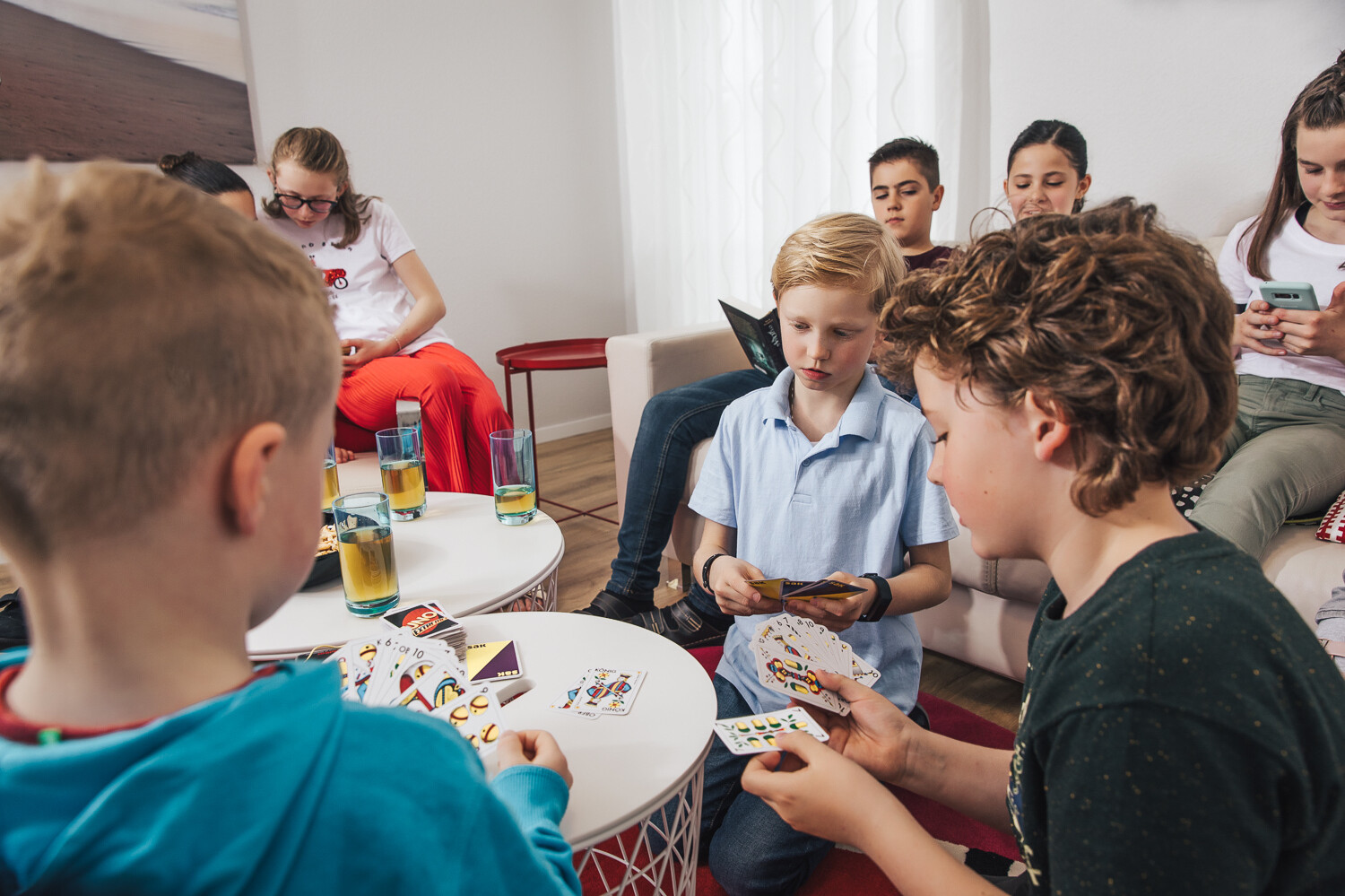Drei Jungen sitzen am Boden des Aufenthaltsraums und jassen mit Karten. Getränke stehen auf dem Tisch neben ihnen. Dahinter sitzen zwei Mädchen, die sich zusammen etwas ansehen. Rechts von den Mädchen sitzt ein Junge auf dem Sofa, der in einem buch liest, während zwei weitere Mädchen rechts von ihm am Handy sind. Die Kindervilla gehört zum Wellnessresort Grand Resort Bad Ragaz, einem Superior Luxusresort in der Schweiz.