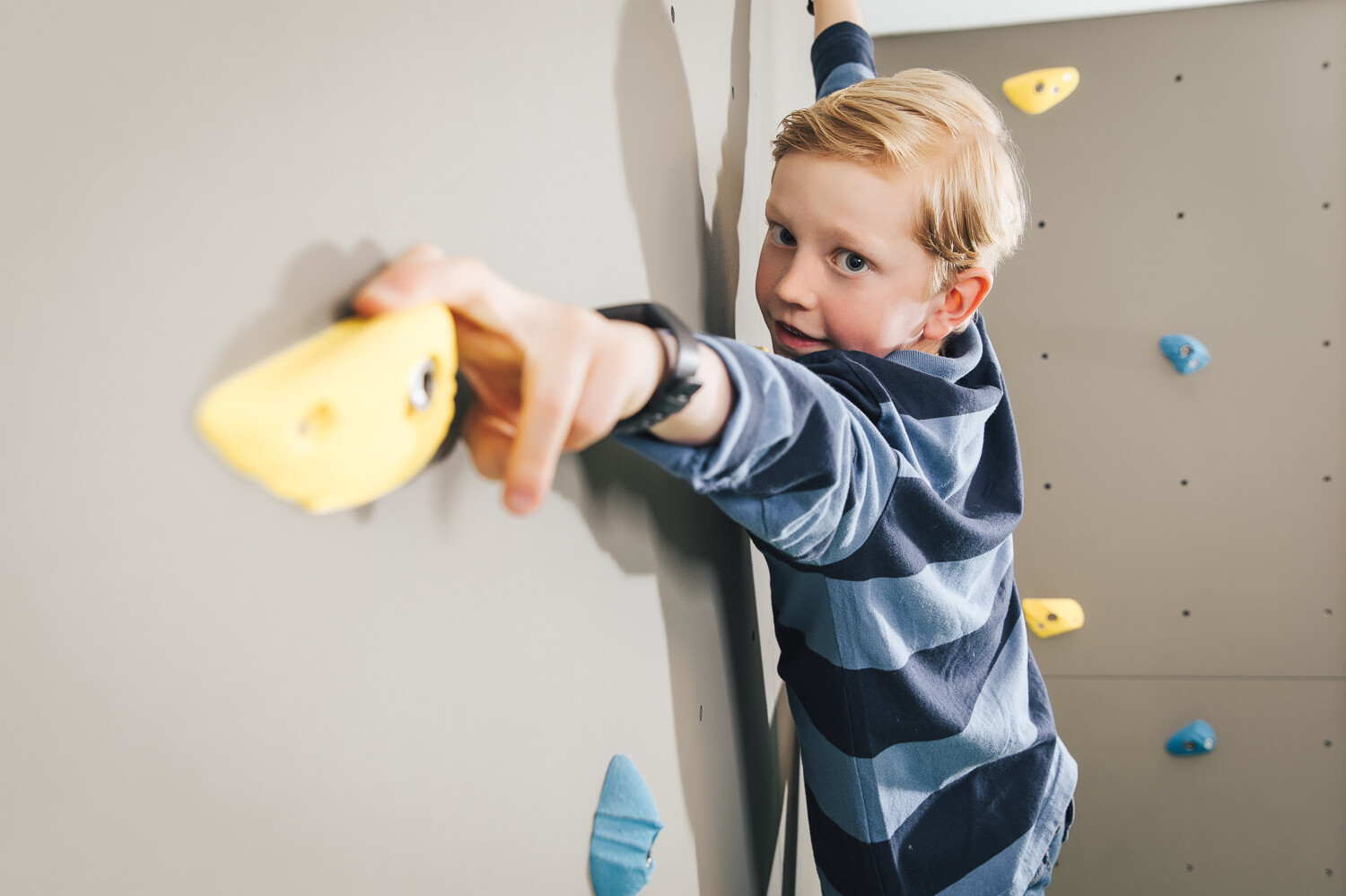 Ein Junge klettert an der Kletterwand in der Kindervilla. Blaue und gelbe Klettergriffe hängen an der grauen Wand. Die Kindervilla gehört zum Wellneshotel Grand Resort Bad Ragaz, einem Superior Luxusresort in der Schweiz.
