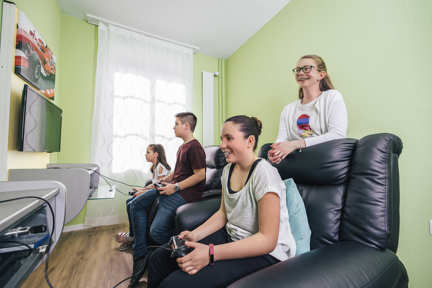 Vier Kinder, drei Mädchen und ein Junge, sitzen in einem grün gestrichenen Raum auf schwarzen Sesseln. Drei der Kinder halten eine Gamingkonsole in der Hand. Das blonde Mädchen ohne Gamingkonsole schaut ihrer Freundin lachend zu. Die Kindervilla gehört zum Wellnessresort Grand Resort Bad Ragaz, einem Superior Luxusresort in der Schweiz.