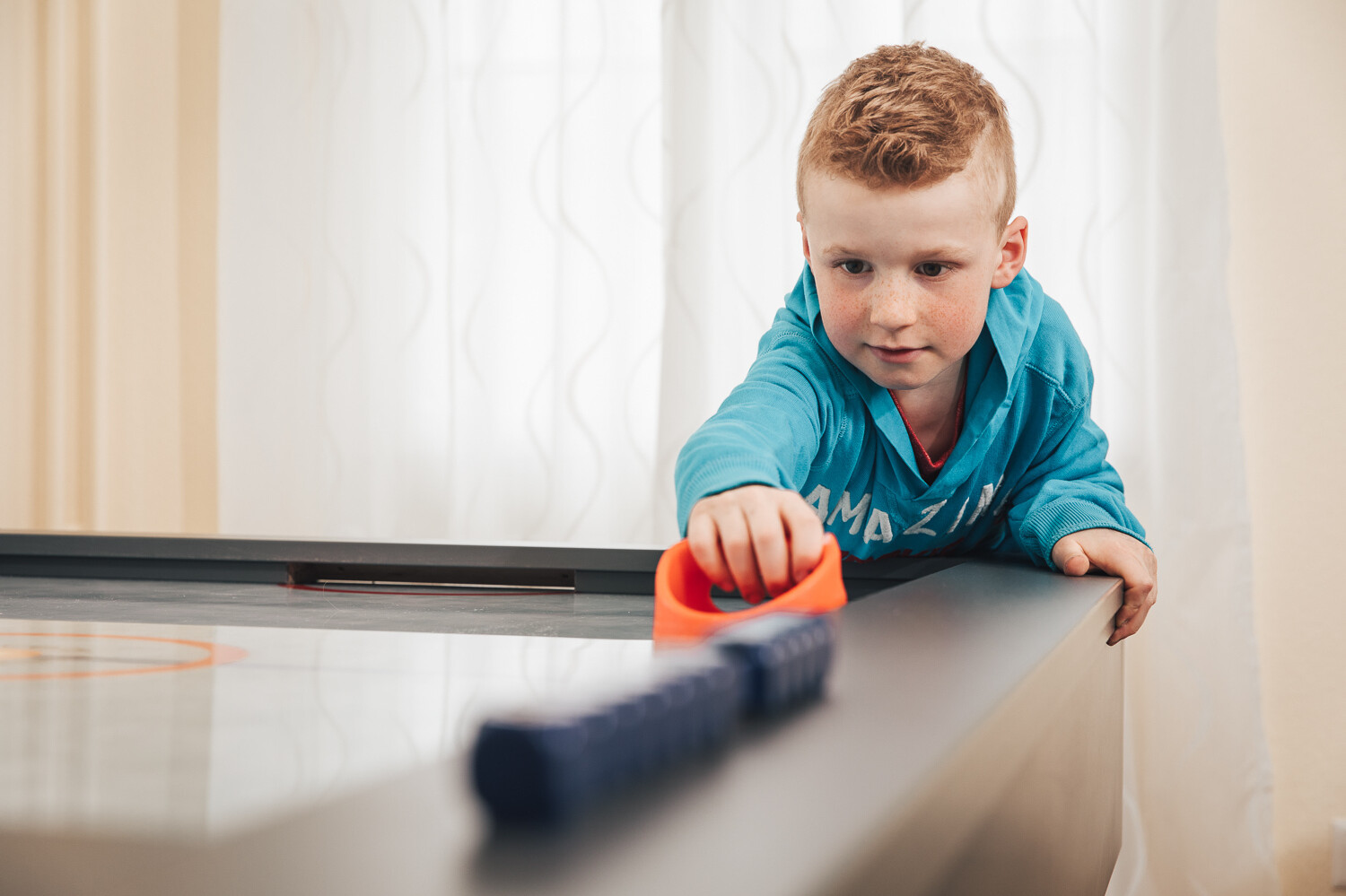Ein kleiner Junge in blauem Pullover mit roten Haaren und Sommersprossen spielt Airhockey mit einem orangnen Plastikteil. Hinter ihm befindet sich ein weisser Vorhang vor dem Fenster. Er befindet sich in der Kindervilla. Diese gehört zu dem Wellnessresort Grand Resort Bad Ragaz, einem Superior Luxushotel in der Schweiz.