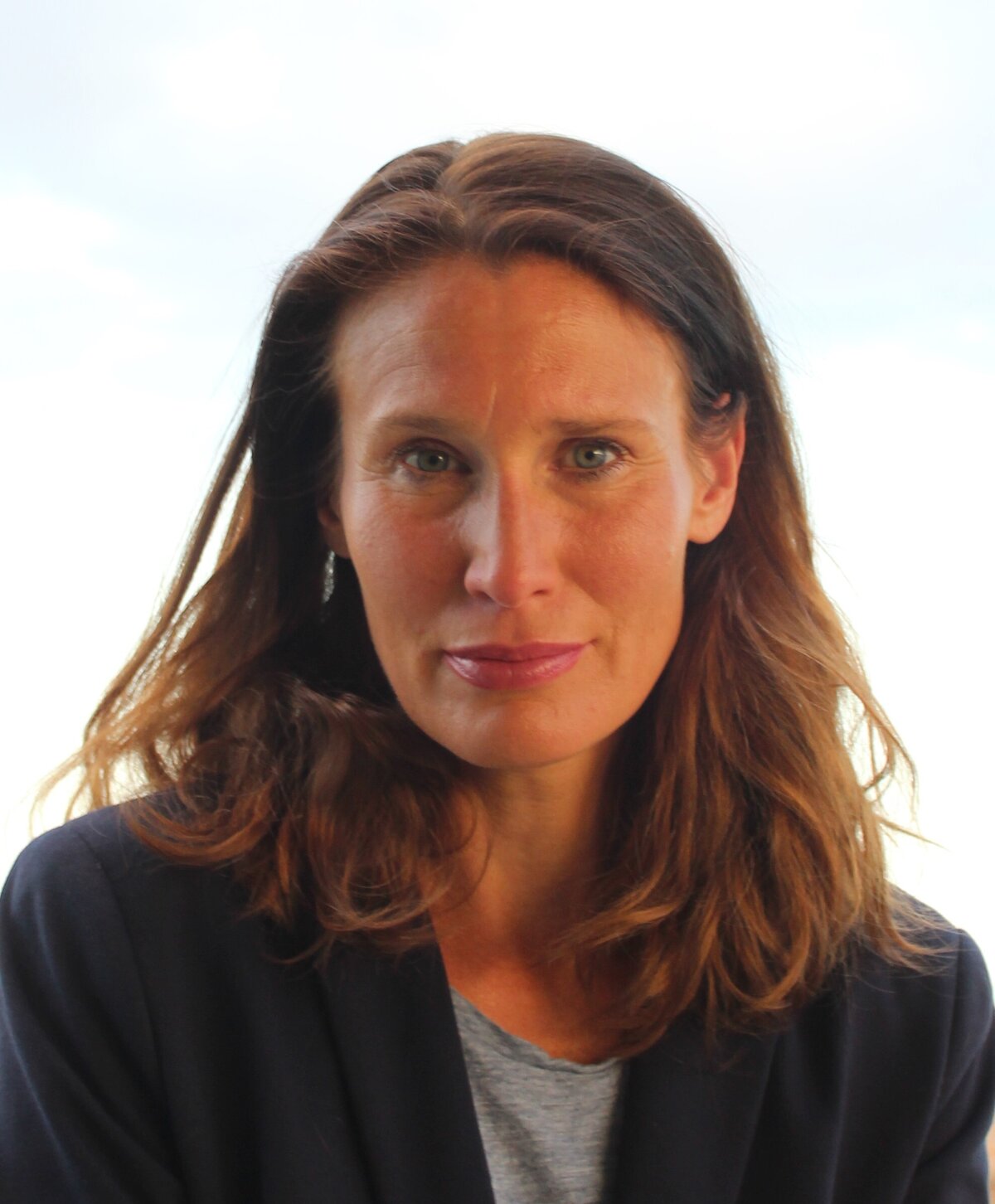 Das gebräunte Gesicht von Corinna Mühlhausen schaut in die Kamera. Ihre braunen Haare fallen bis zu ihren Schultern und ihr blauen Augen strahlen entgegen. Sie trägt einen schwarzen Blazer, der einen starken Kontrast zu dem hellen, weissen Hintergrund bildet.