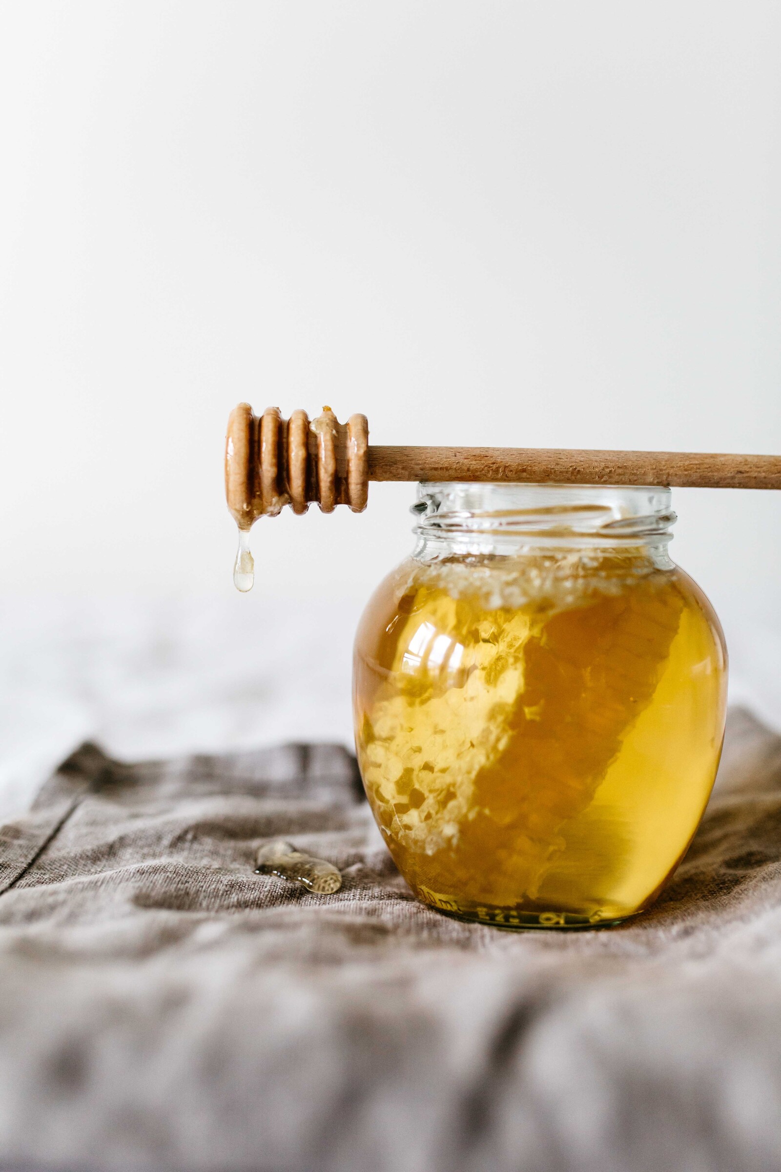 Auf einem grauen Tuch steht ein Glas Honig. Ein kleines Stück der Honigwabe befindet sich in dem Gläschen. Ein Stecken liegt auf dem Glas und Honig tropft von diesem auf das Tuch. Die Wand hinter dem Glas ist weiss.