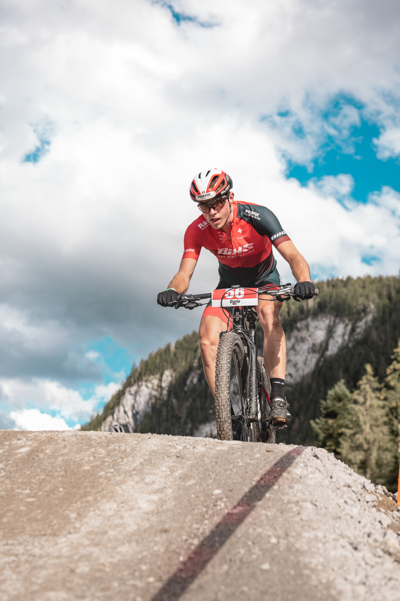 Ein Biker in schwarz-rotem Trainingsanzug biked auf seinem Mountainbike über einen Hügel. Hinter ihm sind die Berge zu sehen. Der Himmel hat ein paar Wolken, aber die Sonne scheint.