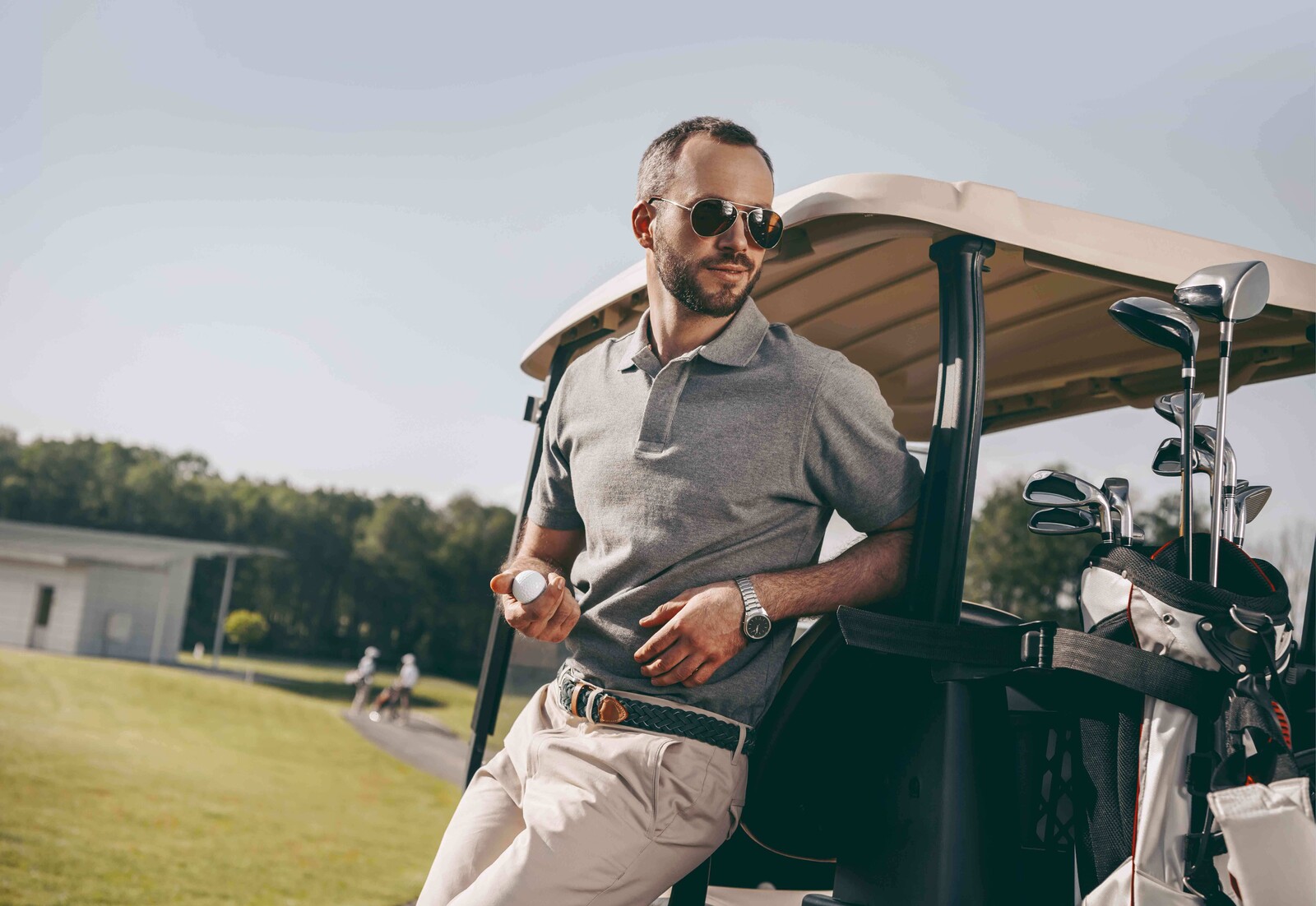 Ein Mann im lässigen Golfoutfit lehnt mit meinem Golfball in der Hand an einen Golf-Caddy.