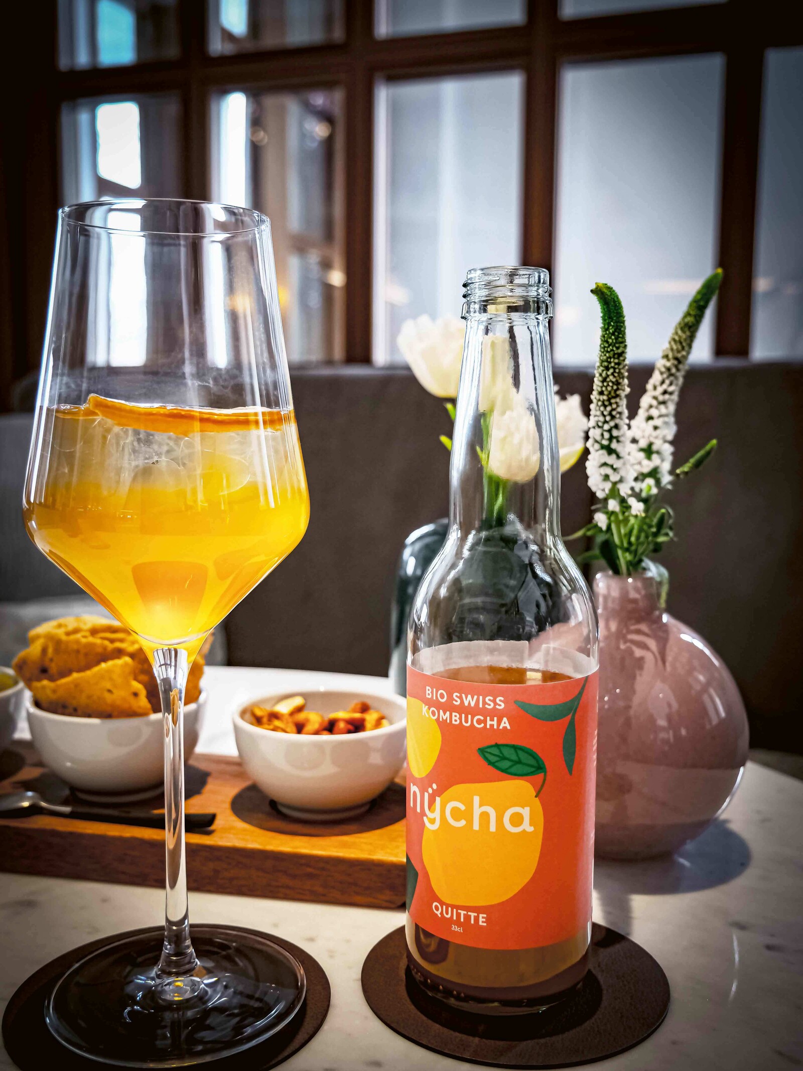 Eine Flasche Kombucha der Marke nycha steht neben einem Glas Kombucha auf dem Tisch. Im Hintergrund sind Snacks und eine Tischdeko platziert.