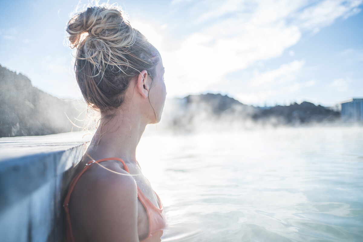 Blick auf das Profil einer jungen Frau, die im Wasser am Beckenrand anlehnt. Aus dem Wasser steigt Nebel auf.
