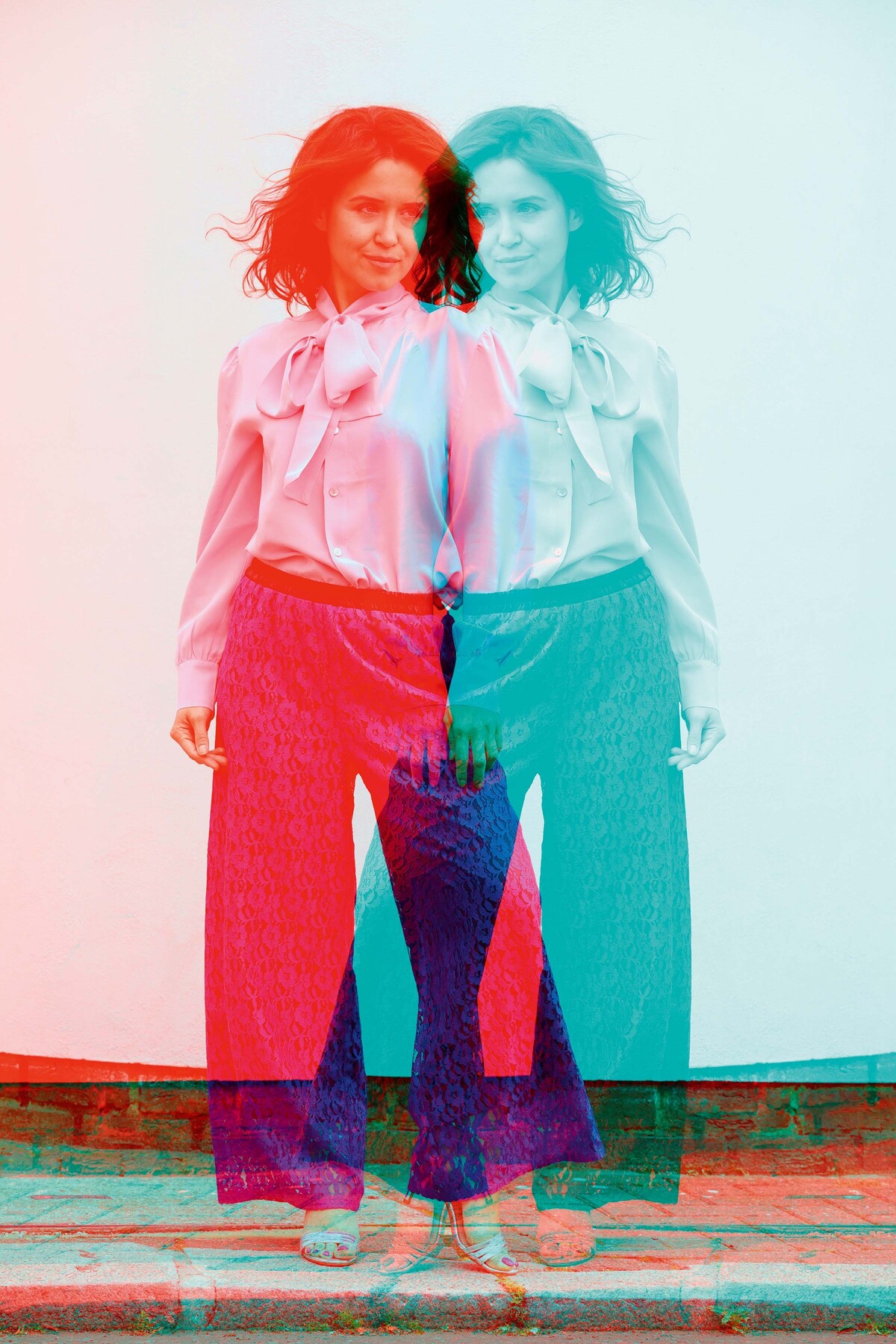 Eine junge Frau wird in diesem Bild gespiegelt. Eine ihrer Versionen ist rot, die andere türkis. Der Betrachter schaut frontal auf die Dame. Sie trägt eine luftige Anzugshose und eine helle Bluse.