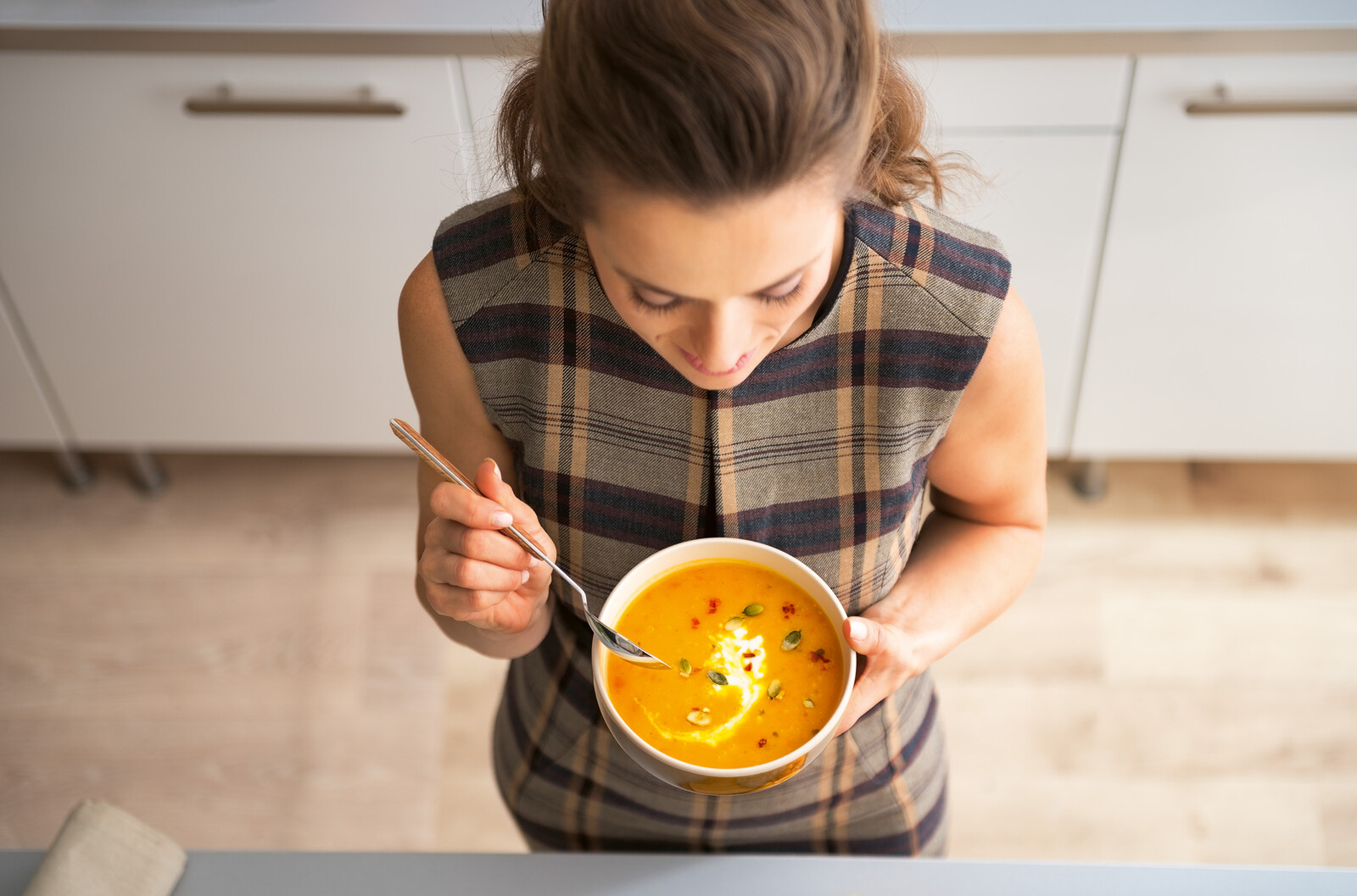 Frontaler Blick auf eine Frau aus der Vogelperspektive. Sie trägt ein herbstliches, kariertes Top und hält eine Schale mit Kürbissuppe und einen Löffel.