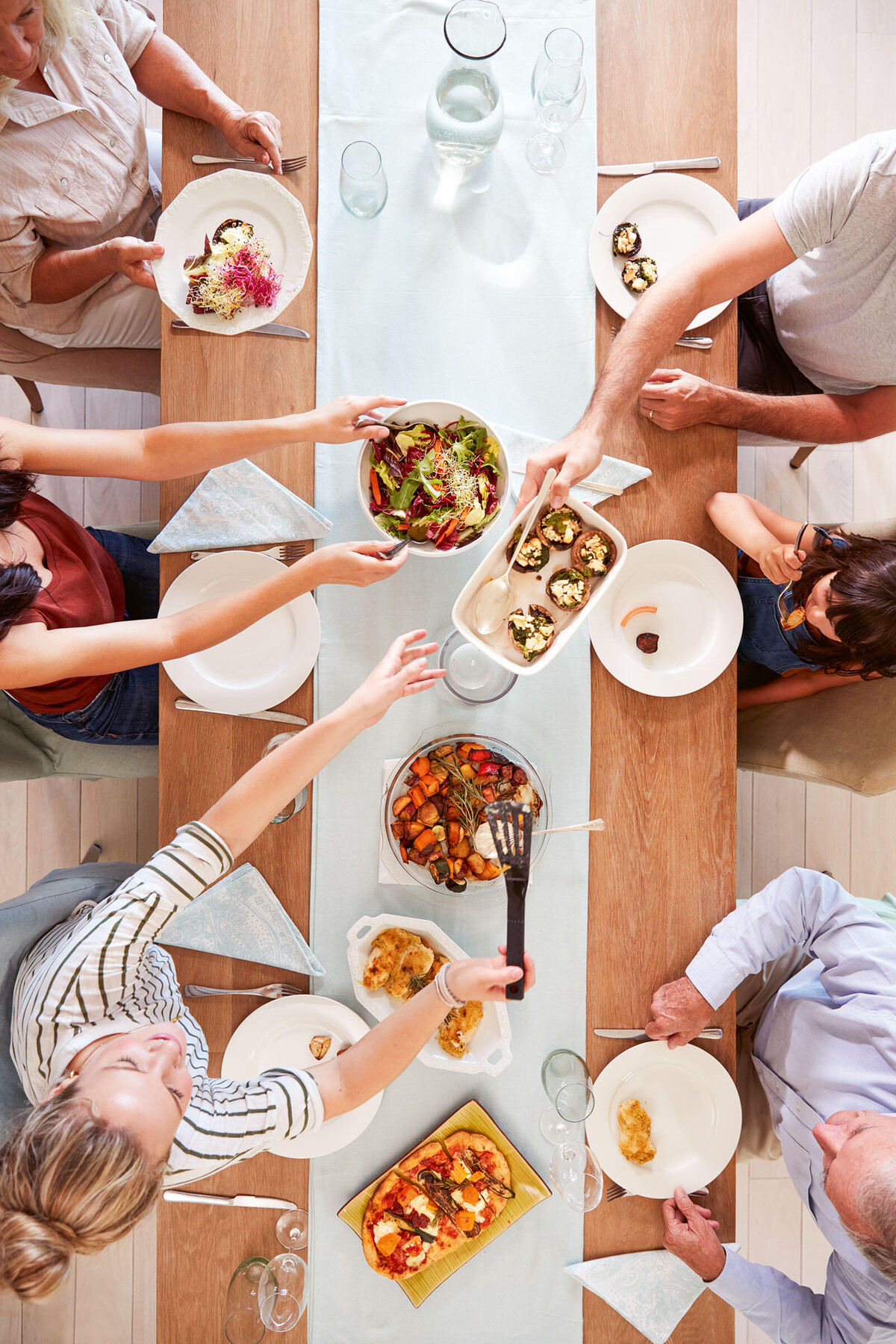 Blick aus der Vogelperspektive auf einen gedeckten Esstisch. Sechs Personen unterschiedlichen Alters sitzen am Tisch, reichen sich Essen und unterhalten sich.