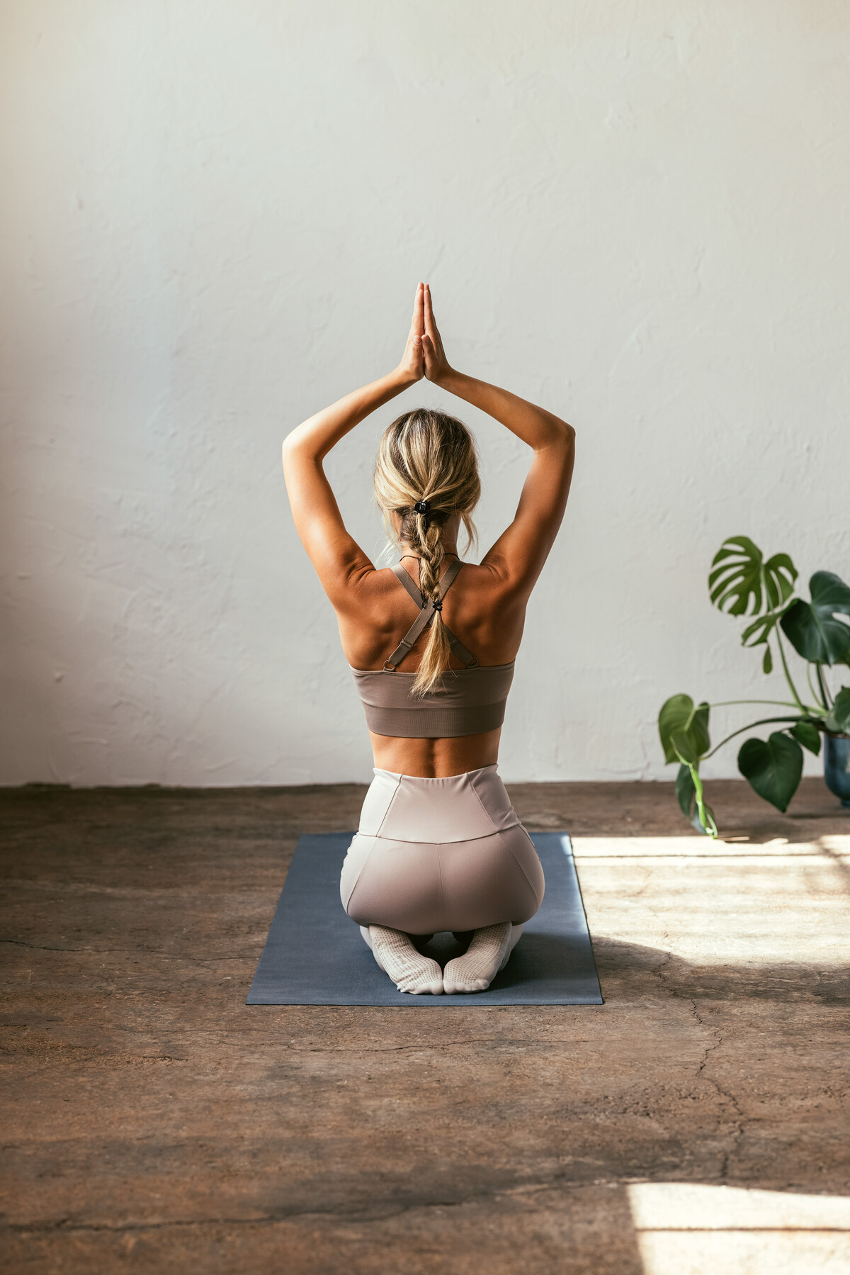 Eine junge Frau in einem beigen Sportoutfit kniet auf einer schwarzen Sportmatte vor einer weissen Wand. Ihre Hände sind über ihrem Kopf zusammengefaltet.