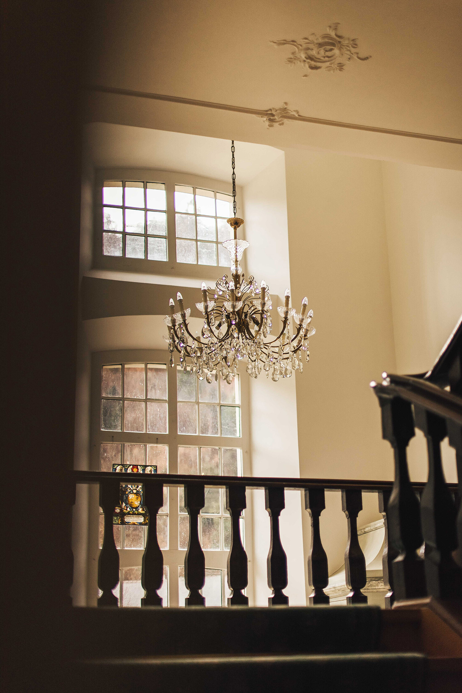 The elegant staircase of the Hotel Palais Bad Ragaz. has wooden railing and a large chandelier in a sunlit room. Tall windows let in warm light, creating a serene atmosphere.