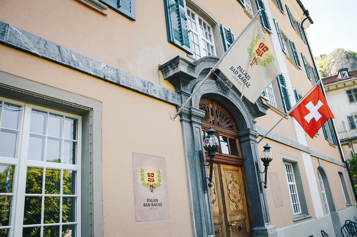 The historic Hotel Palais Bad Ragaz. A beige building with green shutters, featuring a wooden arched door, two flags displayed and decorative lanterns.
