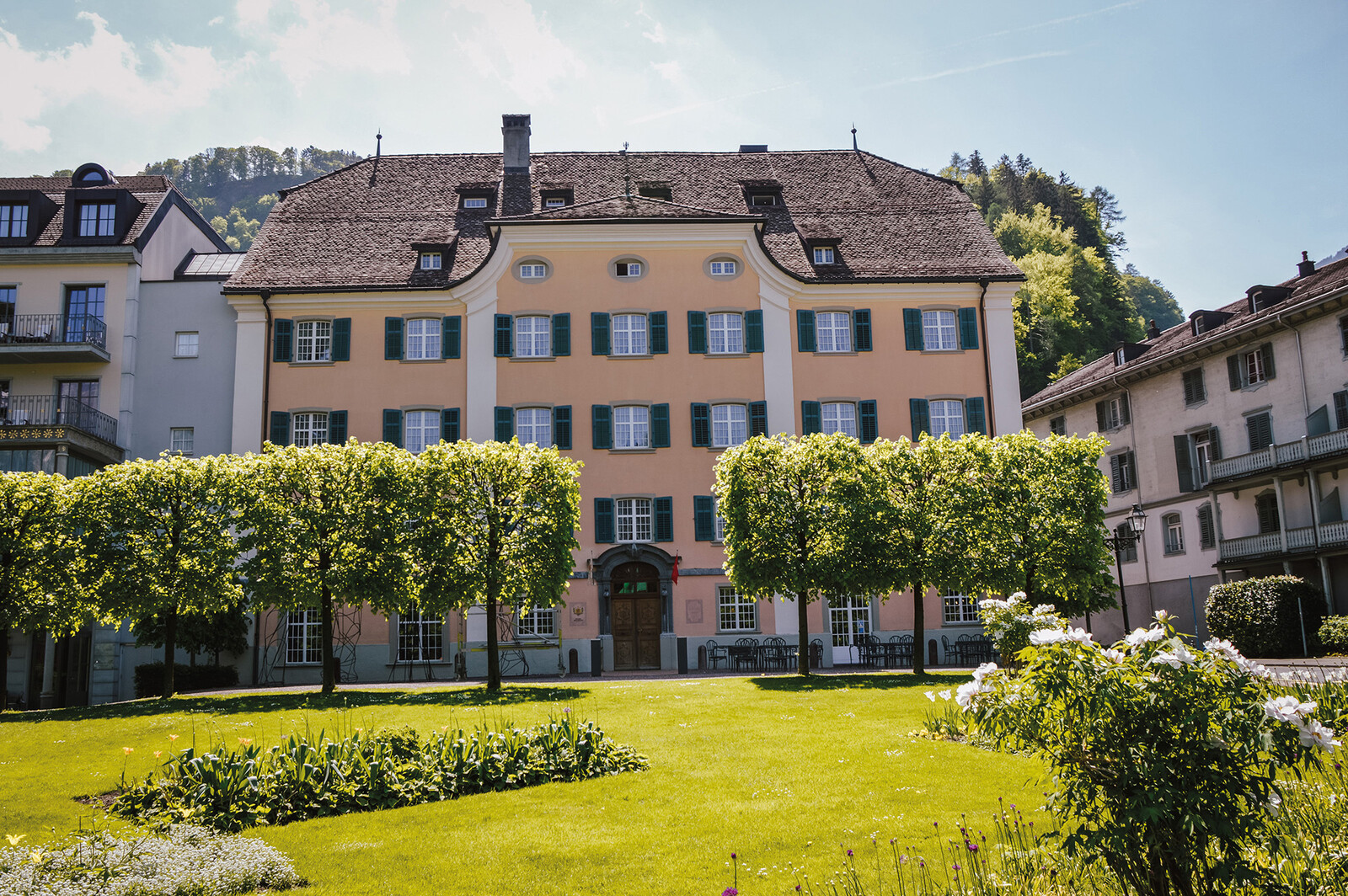 The historic Hotel Palais Bad Ragaz. A beige building with green shutters, framed by trees and lush gardens, under a clear blue sky.