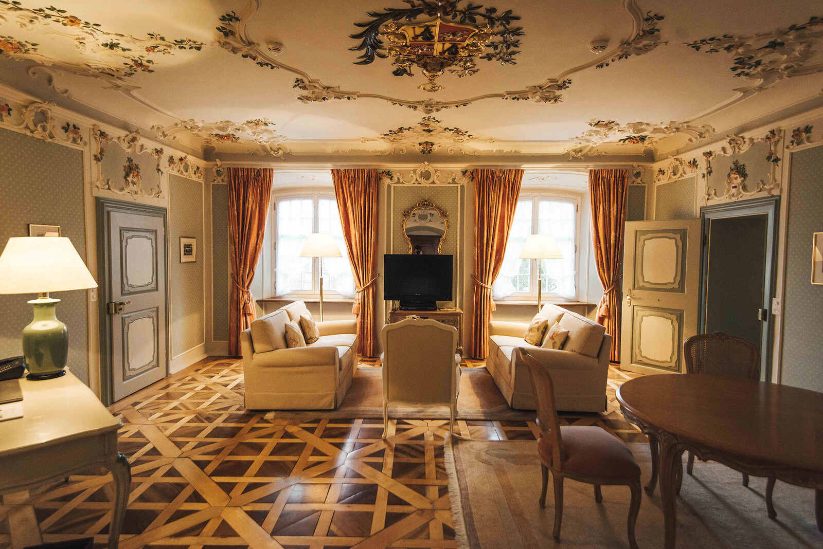 The elegant living room of the Fürstensuite of the Hotel Palais Bad Ragaz. The room has a ornate ceiling, patterned wood floor, and vintage decor featuring beige sofas, a central TV, and large windows with orange drapes.