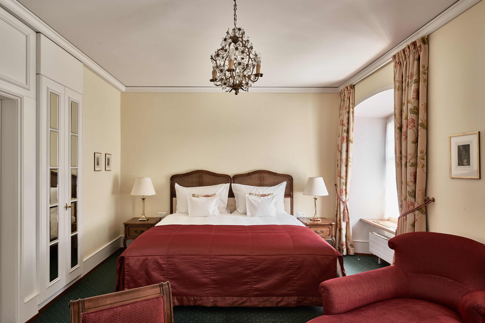 Elegantes Hotelzimmer mit einem plüschigen Doppelbett mit roter Bettwäsche, flankiert von klassischen Holznachttischen. Darüber hängt ein Kronleuchter, und geblümte Vorhänge umrahmen das Fenster und schaffen eine warme, einladende Atmosphäre.