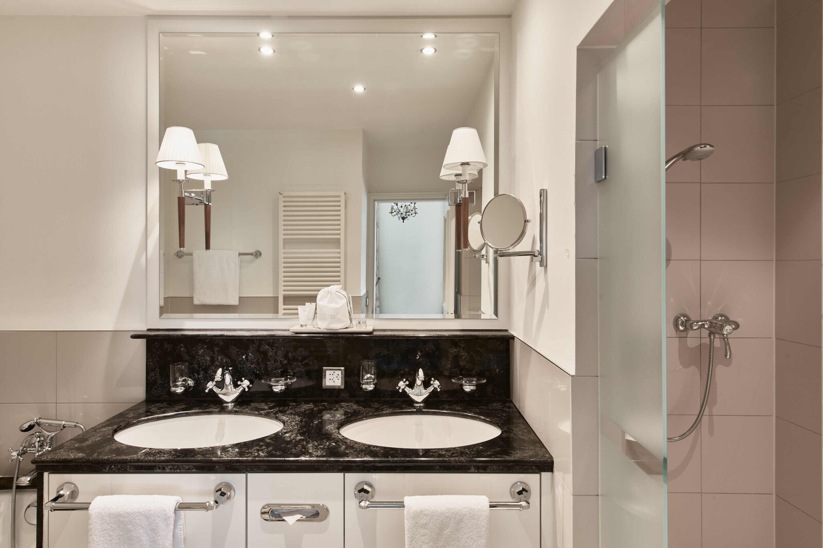 Elegantes Badezimmer mit doppeltem Waschtisch und Arbeitsflächen aus schwarzem Marmor. Zwillingsspiegel, Wandleuchten und eine Dusche mit Glastür sorgen für ein luxuriöses Ambiente.