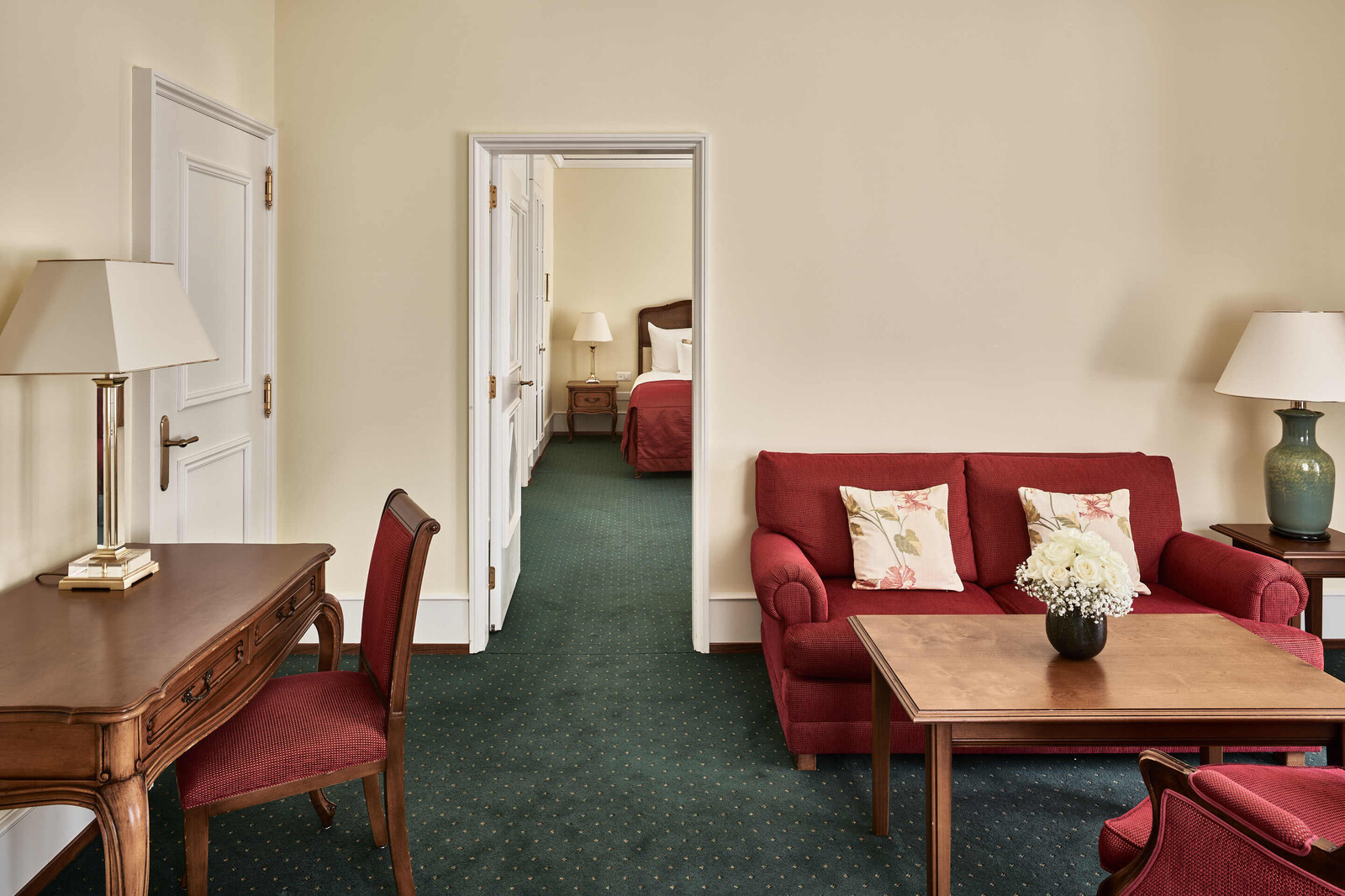 Gemütliches Hotelzimmer mit Vintage-Dekor: rotes Sofa, geblümte Kissen, hölzerner Couchtisch, eleganter Schreibtisch und ein angrenzendes Schlafzimmer mit roter Bettwäsche im Blick.