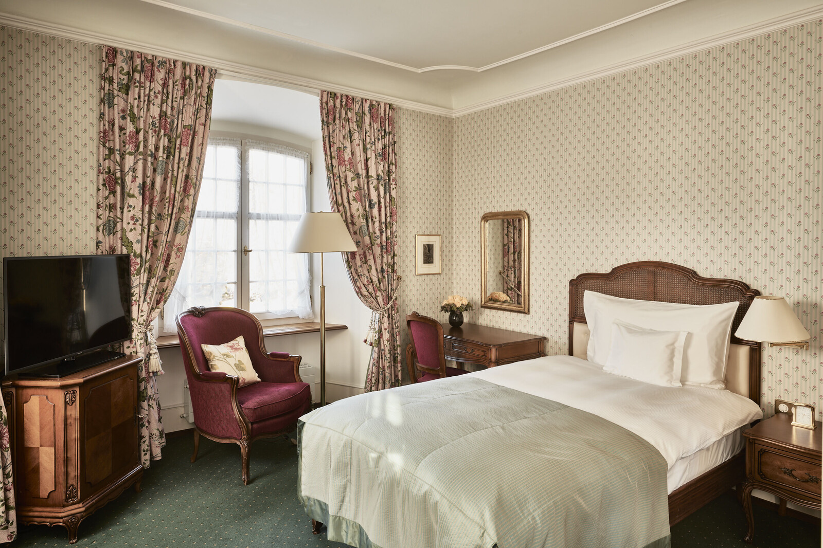 Elegant hotel room with a single bed, antique wooden furniture, floral curtains, and green carpet. The setting is cozy and classic.