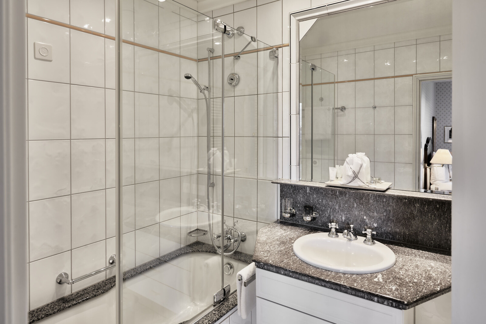 Bright, modern bathroom with white tiled walls. Features a glass-enclosed shower above a bathtub, granite countertop with a sink, and a large mirror.