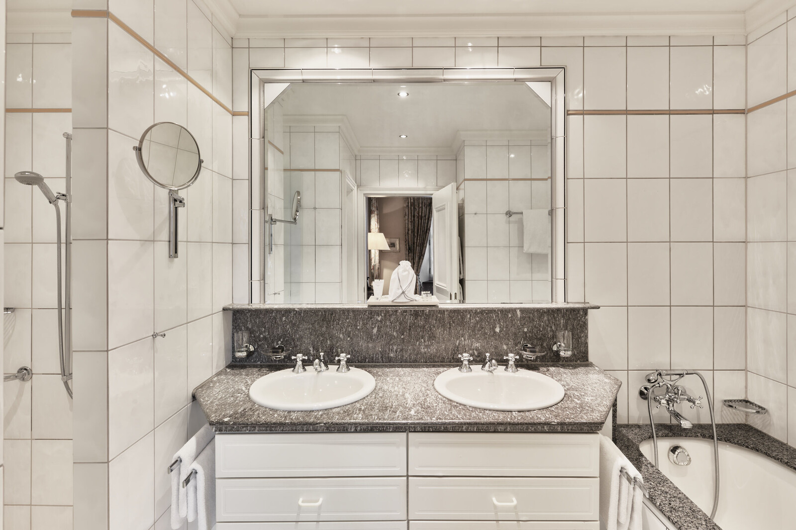 Elegantes Badezimmer mit weiß gefliesten Wänden und einem Doppelwaschbecken mit einem großen Spiegel. Auf der rechten Seite befindet sich eine Badewanne, und es gibt eine warme, stimmungsvolle Beleuchtung.