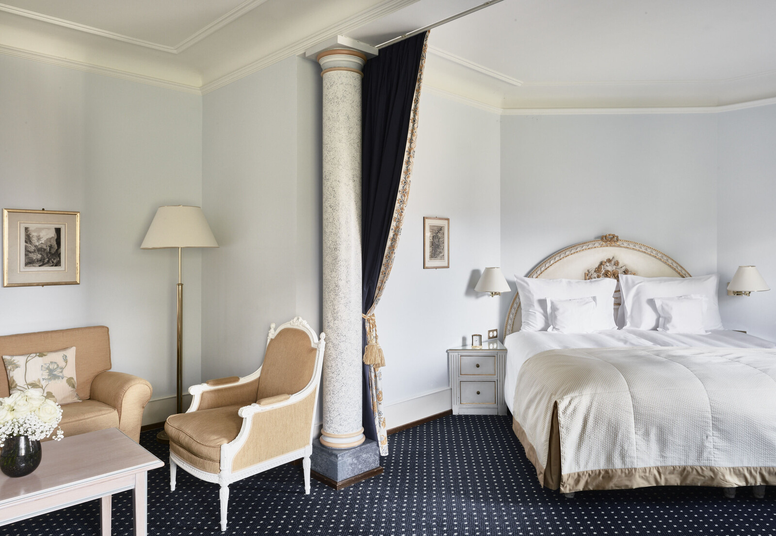Elegantes Hotelzimmer mit plüschigem beigem Sessel, passendem Sofa und blumengeschmücktem Tisch. Ein großes Bett mit verschnörkeltem Kopfteil und blauen Vorhängen strahlt Luxus aus.