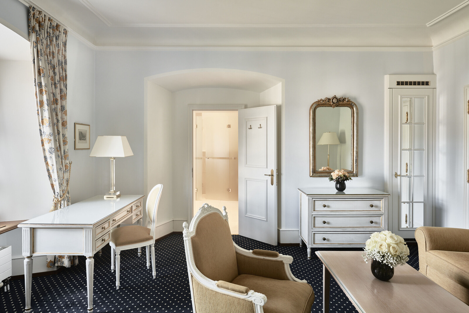 Elegantes Zimmer mit Vintage-Dekor: weisser Schreibtisch, Stühle, Kommode mit Spiegel, geblümter Vorhang und eine Vase mit weißen Blumen auf dem Couchtisch.