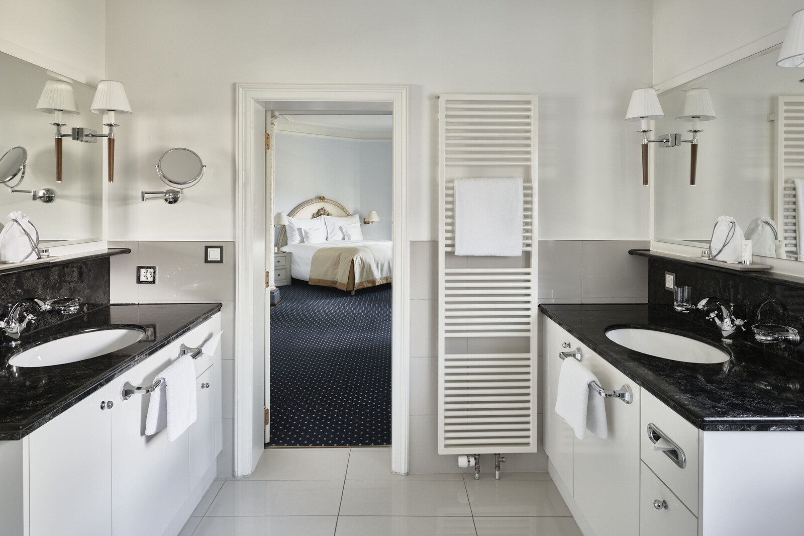 Elegantes Badezimmer mit zwei schwarzen Granitwaschbecken, weißen Schränken und Wänden. Eine Tür öffnet sich zu einem gemütlichen Schlafzimmer mit einem Plüschbett, das ein ruhiges Ambiente schafft.