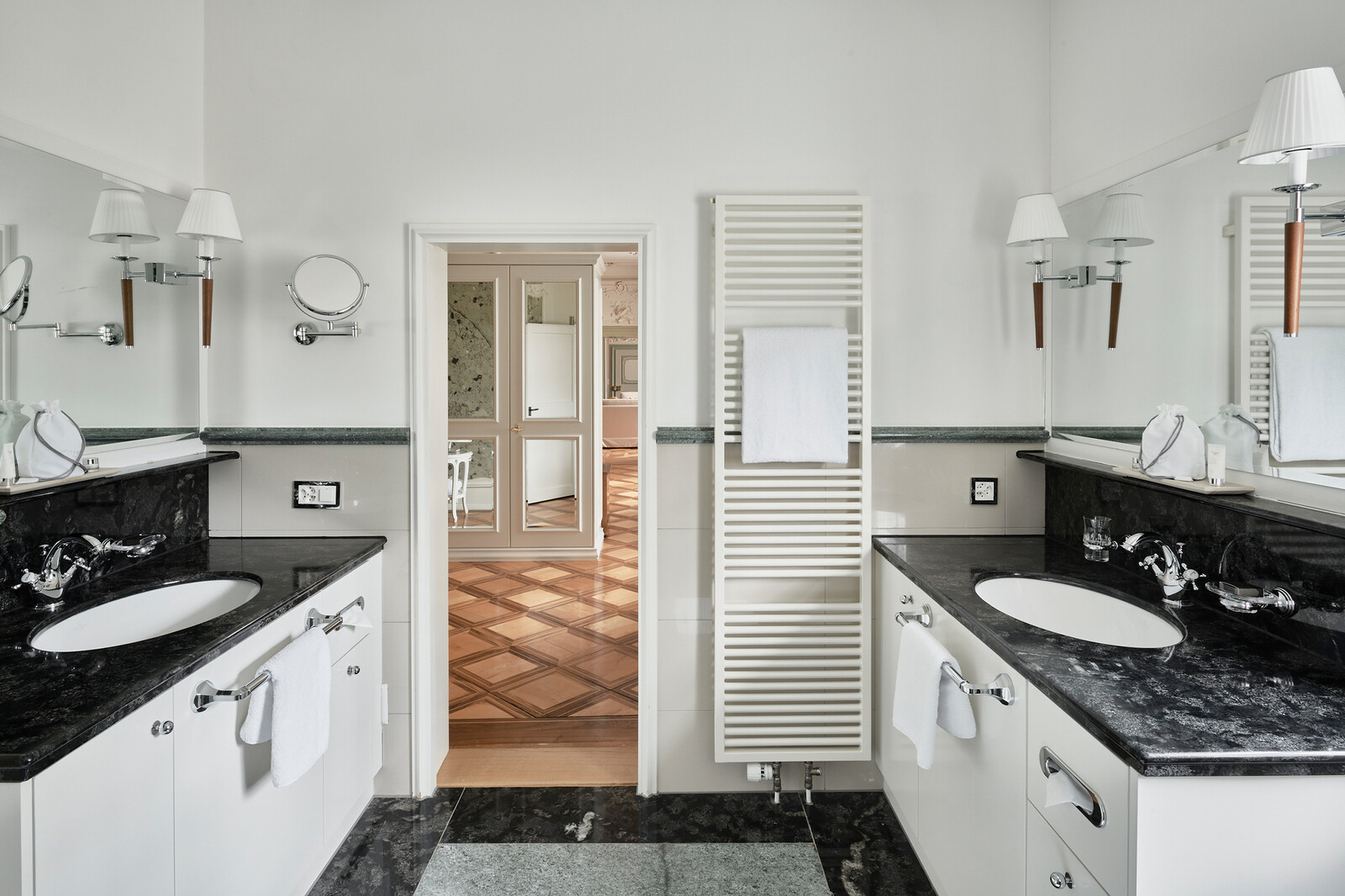 Elegantes Badezimmer mit Arbeitsflächen aus schwarzem Marmor, Doppelwaschbecken und großen Spiegeln. Weisse Wände, Handtuchhalter und eine offene Tür geben den Blick auf einen Flur mit Parkettboden frei.