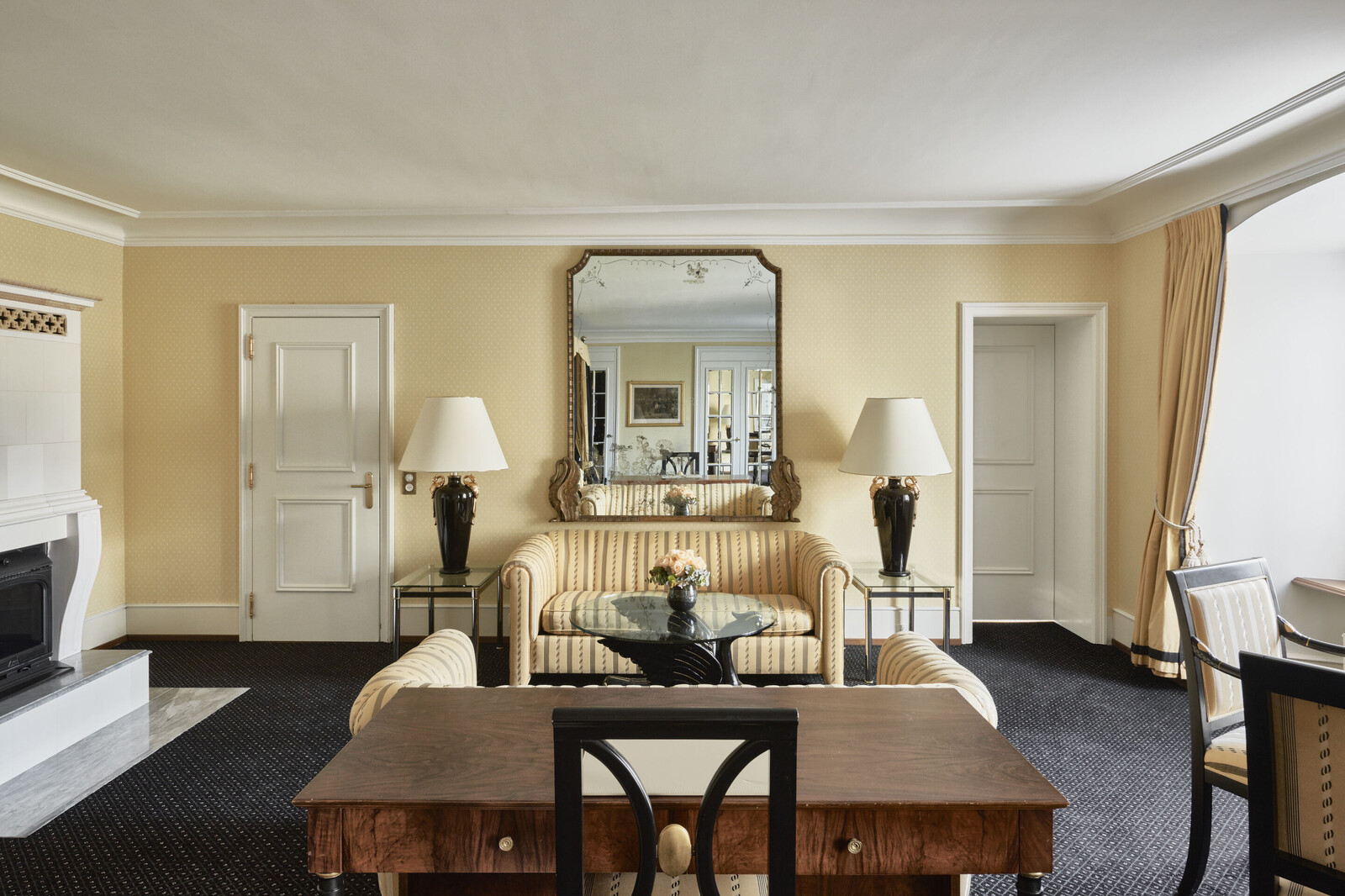 Elegant hotel room with beige walls and a large mirror above a striped sofa, flanked by black lamps. A wooden desk faces a cozy, inviting fireplace.
