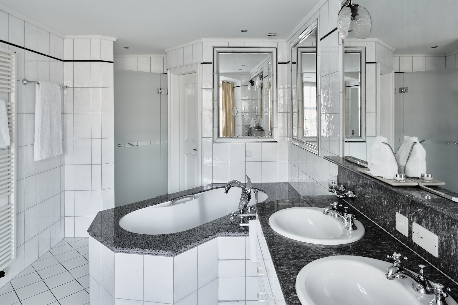 Modern bathroom with white tiled walls and floor. Features double sinks with chrome faucets on a dark countertop, a large bathtub, mirrors, and soft lighting.