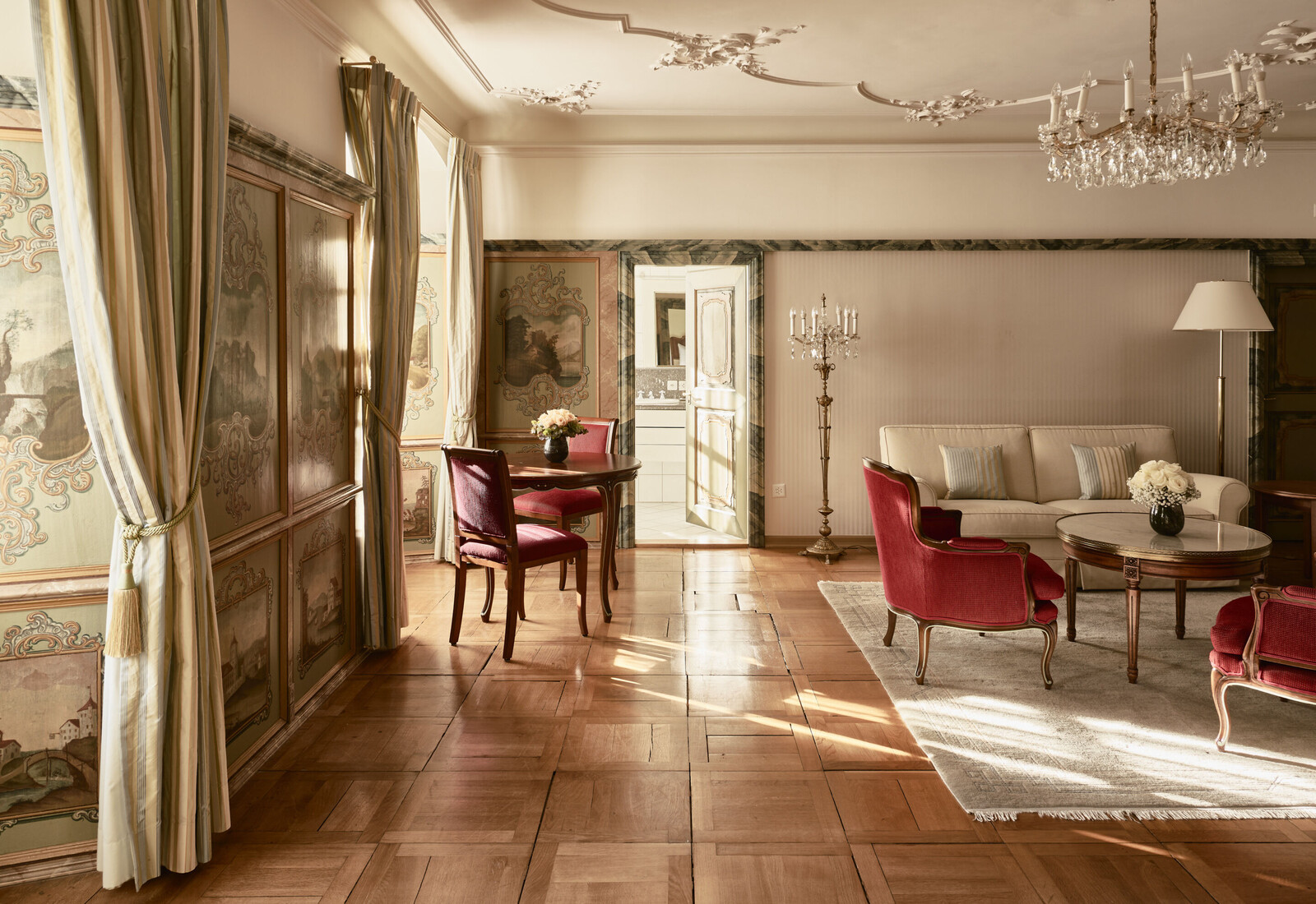 The elegant living room of the Fürstensuite in the Hotel Palais Bad Ragaz. The room has ornate decor, featuring a cream sofa, red velvet chairs, chandelier, and large windows with curtains.