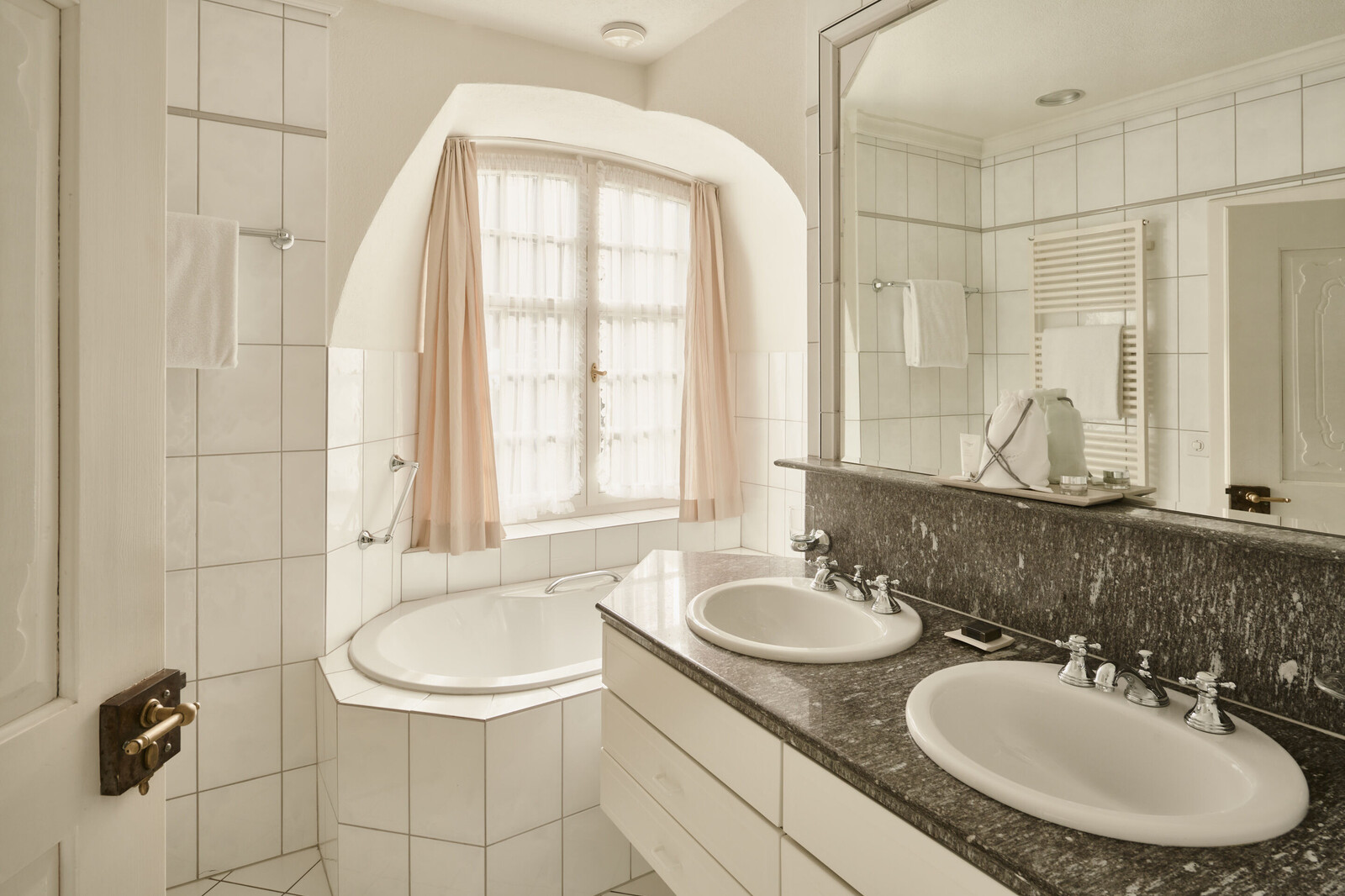Elegant bathroom with white tiles, a large mirror, granite countertop, double sinks, and a corner bathtub. Soft lighting creates a serene atmosphere.