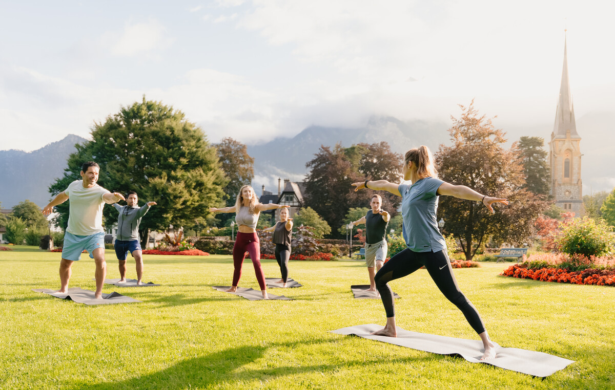 Eine Gruppe von Menschen praktiziert Yoga im Freien auf einer Wiese vor einer Bergkulisse. Der Yogalehrer führt sie in die Kriegerpose.