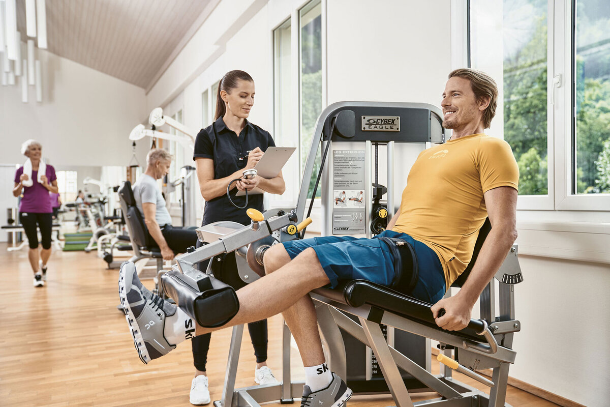 Ein Mann in einem gelben Hemd trainiert lächelnd an einer Beinpressmaschine, während ein Trainer mit einem Klemmbrett ihm dabei hilft. Andere trainieren im Hintergrund. Helles, luftiges Fitnessstudio.