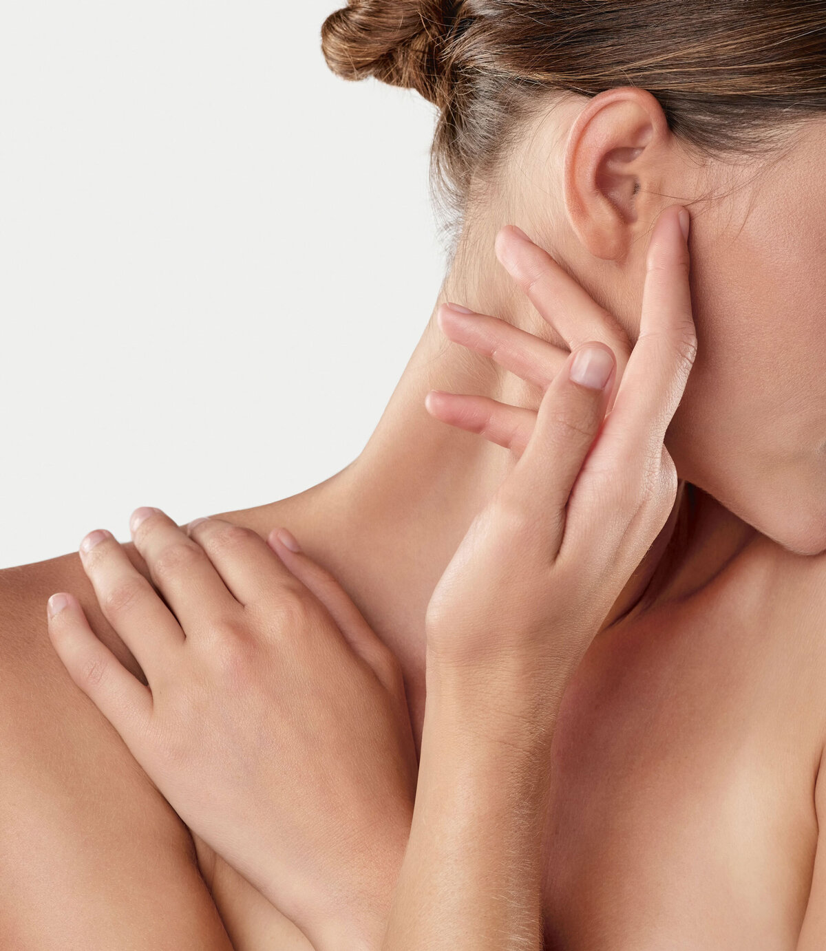 Eine Frau berührt sanft ihr Ohr und ihre Schulter und vermittelt so ein Gefühl der Ruhe und Entspannung. Der Fokus liegt auf ihren Händen und ihrer glatten Haut, der Hintergrund ist neutral.