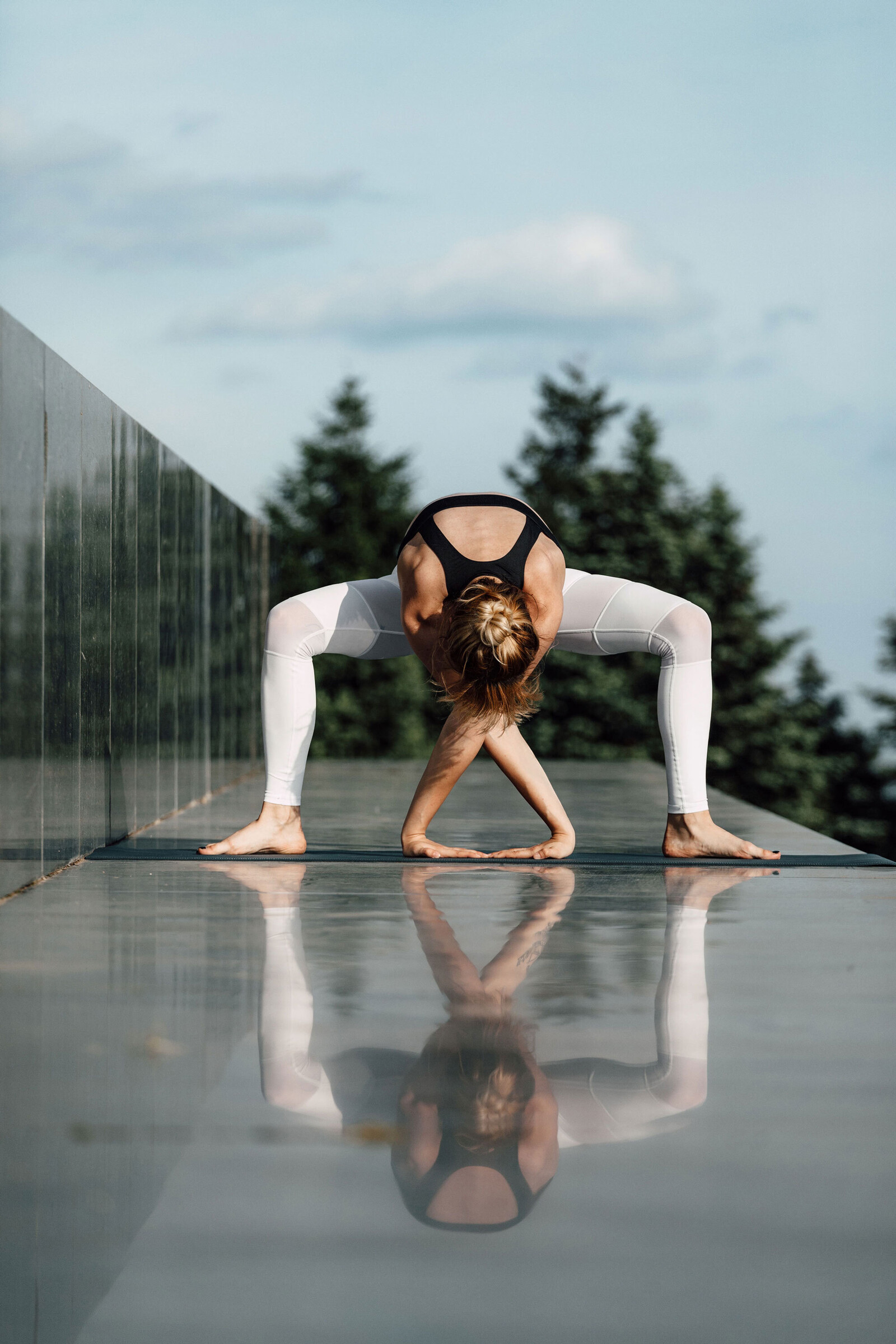 Eine Frau in einem schwarz-weissen Yoga-Outfit macht eine Vorwärtsbeuge auf einer reflektierenden Oberfläche im Freien, mit Bäumen und einem klaren Himmel im Hintergrund.