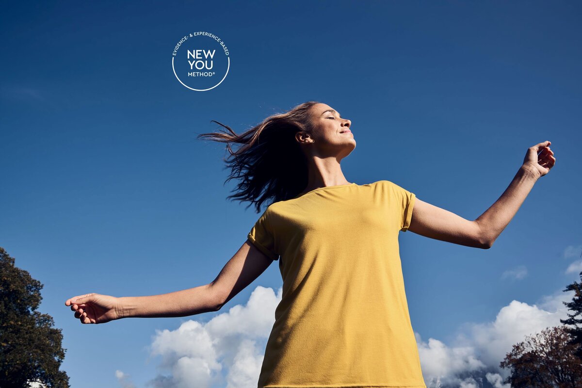 Eine Frau in einem gelben Hemd mit freudig ausgebreiteten Armen vor einem blauen Himmel. Bäume und Wolken im Hintergrund.